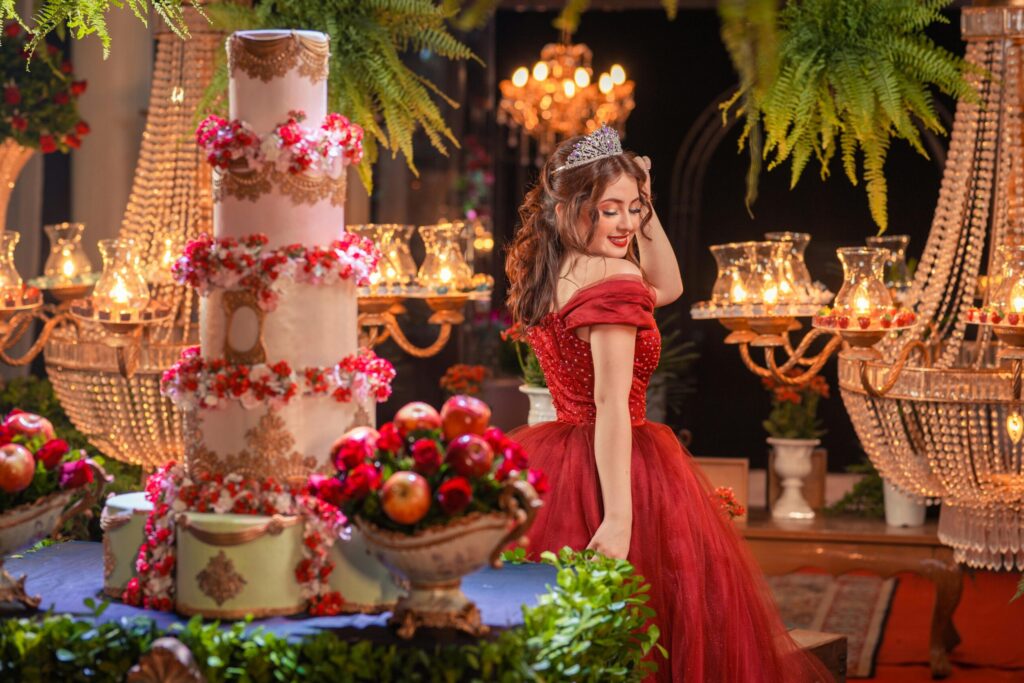 A glamorous woman in a red dress poses beside a grand tiered cake at an elegant indoor celebration.